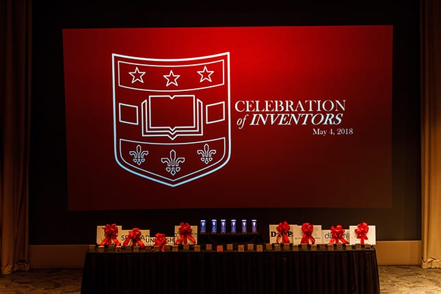 Image of table with various awards; in background is a large projection screen with Washington University shield logo and text reading "Celebration of Inventors, May 4, 2018".